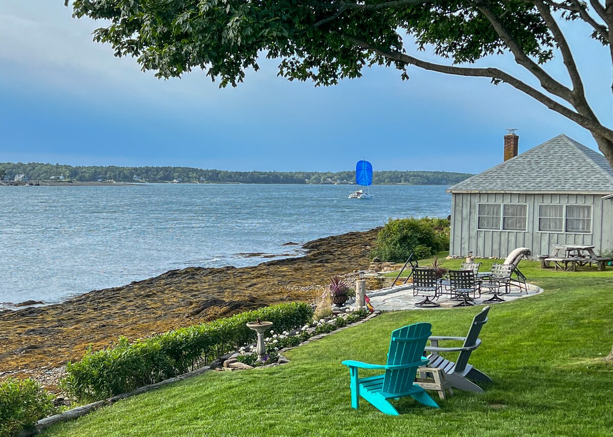 A view of Merriconeag Sound from the yard features a patch of green lawn leading to rocky waterfront. Adirondack chairs in vibrant colors are arranged on the grass, while a small seating area with loungers is visible near the water's edge.
