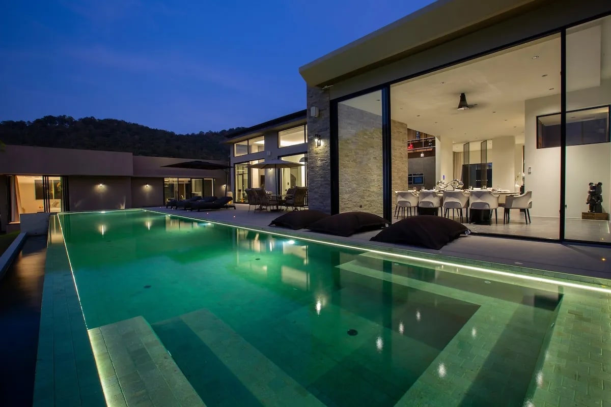 The serene infinity saltwater pool reflects the dusk sky, bordered by comfortable loungers. The villa's modern architecture is showcased, with expansive glass doors inviting light into the open living space. A stylish dining area is visible inside, creating a harmonious indoor-outdoor connection.