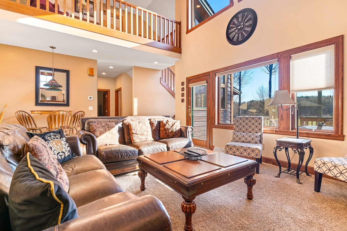 The living room features comfortable seating arrangements, including a spacious leather couch and patterned armchairs. A wooden coffee table rests in the center, surrounded by large windows that invite natural light and offer views of the outdoor scenery. A staircase leads to an upper level.