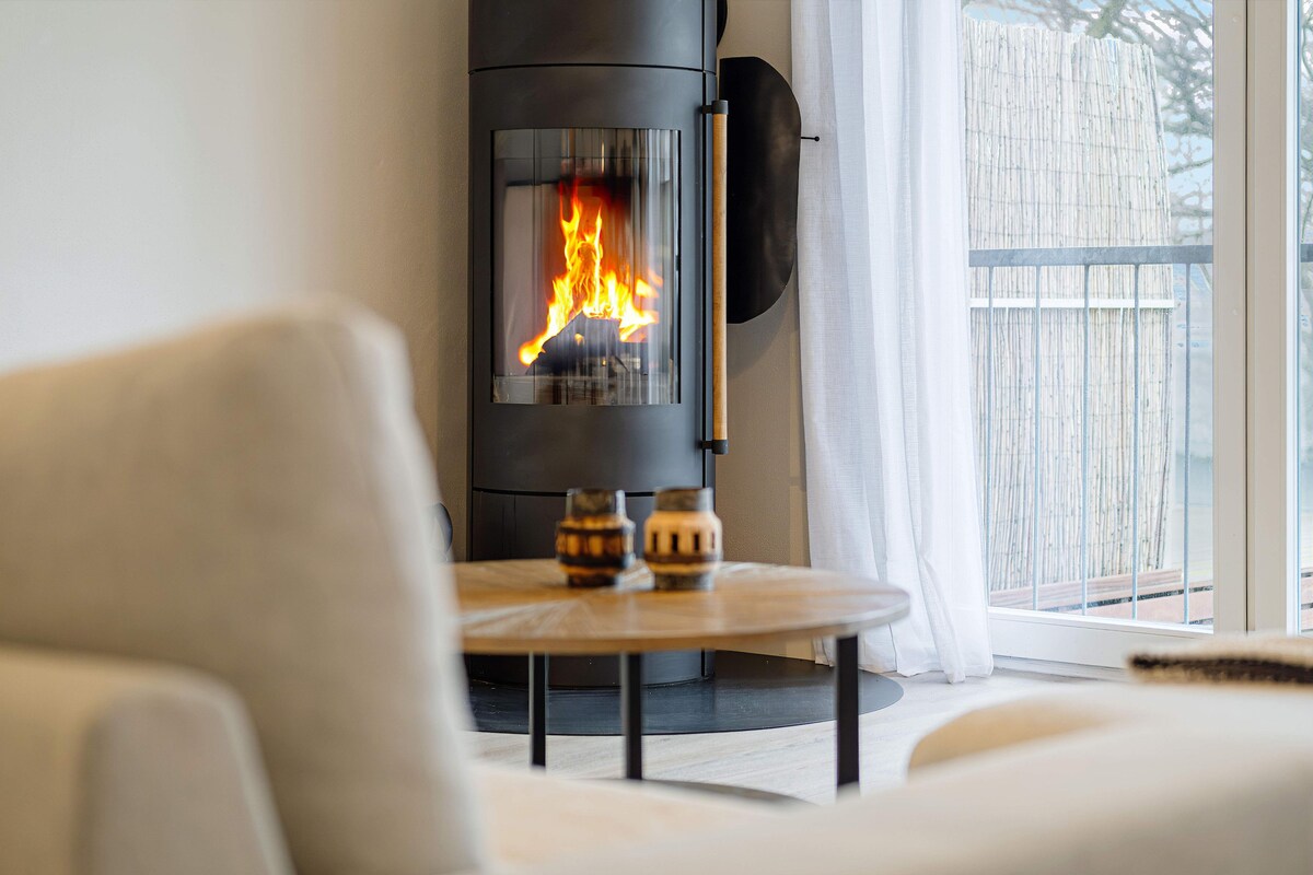 A modern wood stove is seen glowing with flickering flames, positioned beside a large window draped in sheer curtains. A round table made of light wood sits centrally in front of a cozy seating area, enhancing the inviting atmosphere.