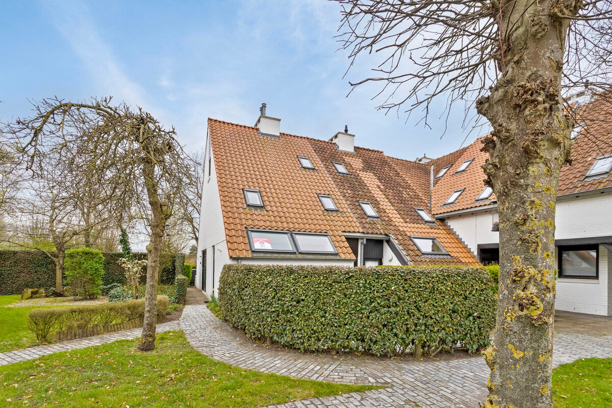 The exterior of the holiday home is shown, featuring a sloped roof with multiple skylights. The structure is surrounded by well-maintained greenery and a brick pathway leads to the entrance. Nearby trees provide natural shade in the serene outdoor setting.