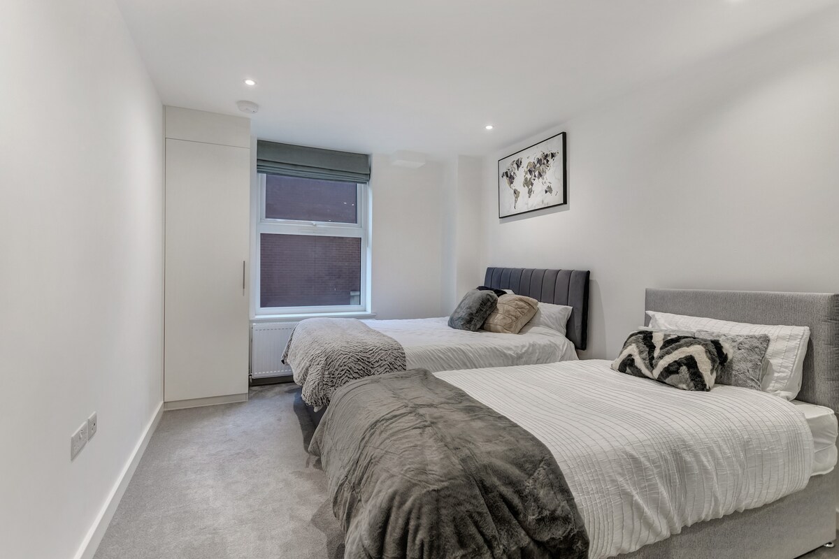 A well-lit bedroom features two beds, each with neutral-toned linens. A window allows natural light to fill the room, while soft carpets add comfort underfoot. Artwork is displayed on the wall above one of the beds, enhancing the room's modern design.