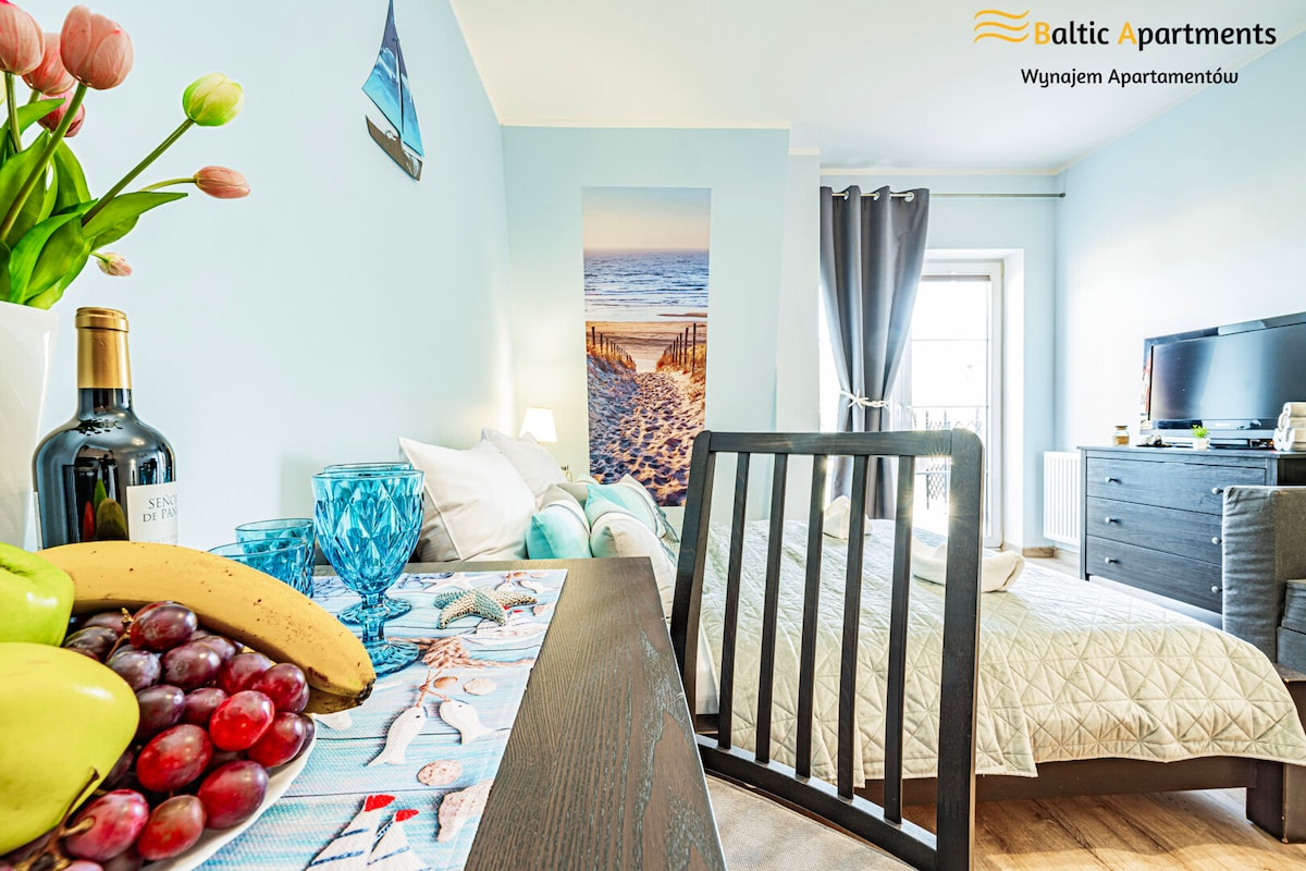 A well-lit room features a double bed with light bedding and a single bed nearby. A dining table with a fresh fruit arrangement sits in the foreground, while a decorative beach-themed wall art adds a coastal touch. Natural light fills the space through a window with gray curtains.