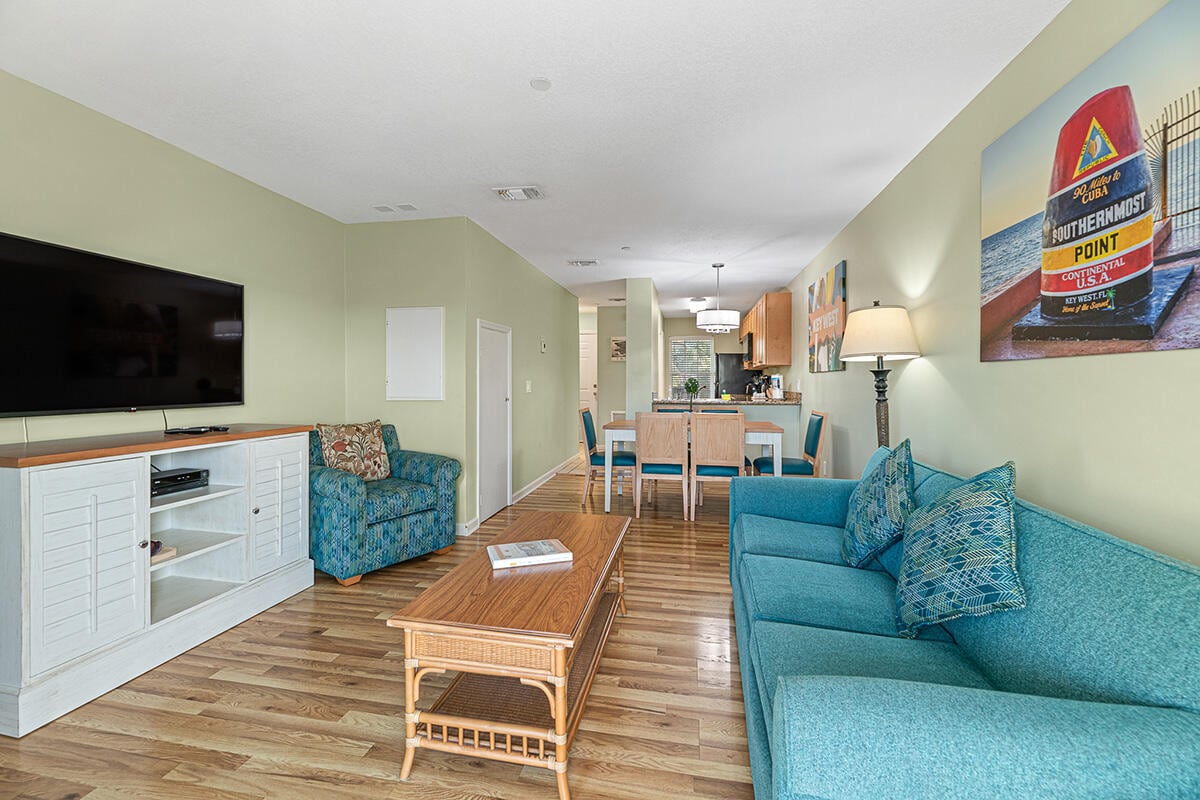 A spacious living area features a turquoise couch and an armchair with a patterned fabric. A wooden coffee table sits centered on the hardwood floor. A large flatscreen TV is mounted on the wall, while a dining area with a table for six can be seen in the background.