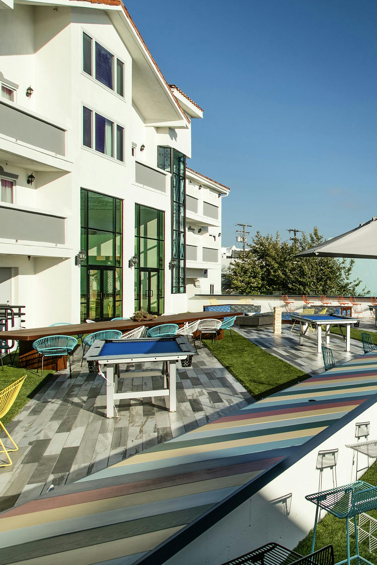An inviting outdoor patio area features colorful furniture, including chairs and tables, arranged for gathering. A lively atmosphere is created by the lush green grass and sun-drenched deck, while expansive glass doors lead into the hotel building, enhancing the connection between indoor and outdoor spaces.