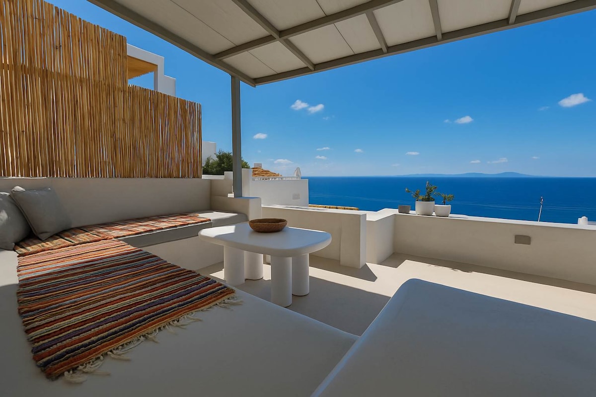A spacious private balcony features comfortable seating and a low table, offering stunning views of the blue sea. Natural light filters through, enhancing the serene atmosphere. Decorative elements include a striped outdoor rug and potted plants, creating a cozy outdoor retreat.