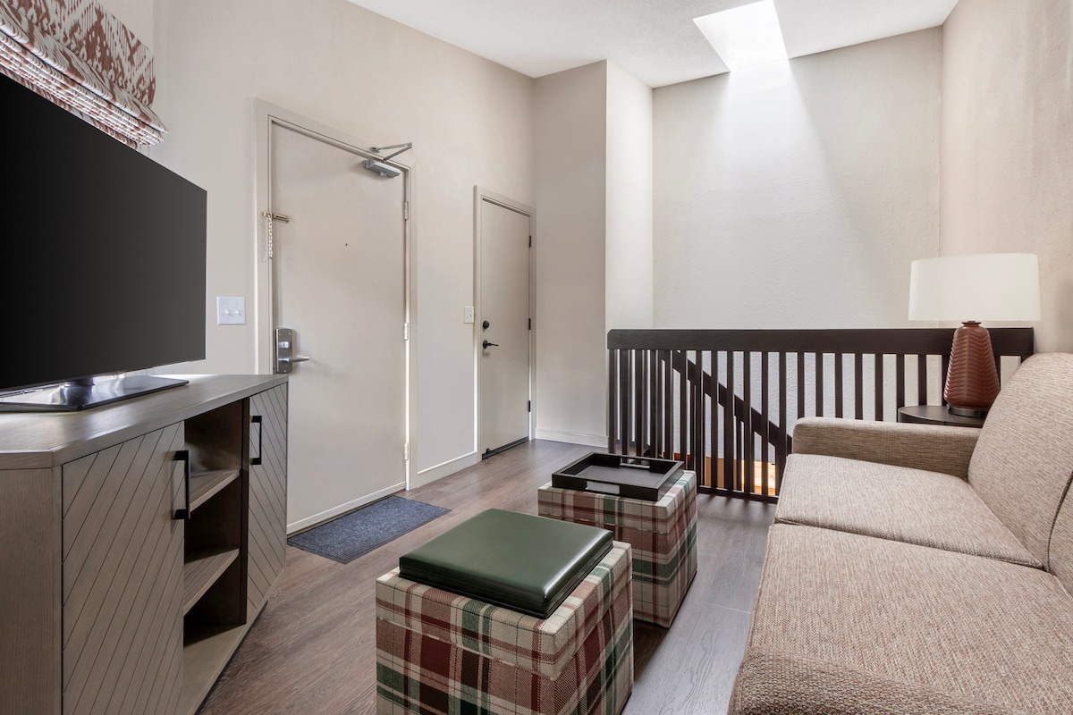 A bright entryway features a cozy seating area with a textured sofa and plaid ottomans. Natural light filters through a skylight, illuminating the space. The room includes a console with a television and provides easy access to the stairway.