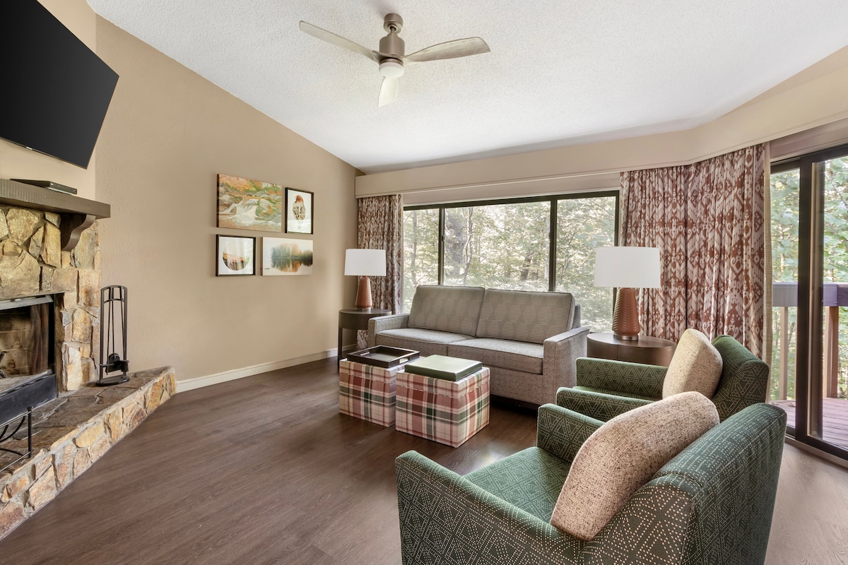 The living area features a stone fireplace and large windows that provide natural light and views of the surrounding nature. A comfortable sofa is complemented by two patterned chairs and a plaid coffee table. Artwork adorns the walls, enhancing the cozy ambiance.