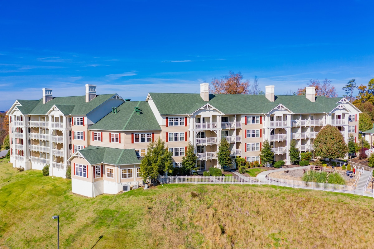The multi-story building showcases a blend of modern and traditional architecture, featuring a green roof and pale facade. Well-maintained landscaping surrounds the property, with neatly arranged trees and shrubs. A patio area is visible, offering space for outdoor seating and relaxation.