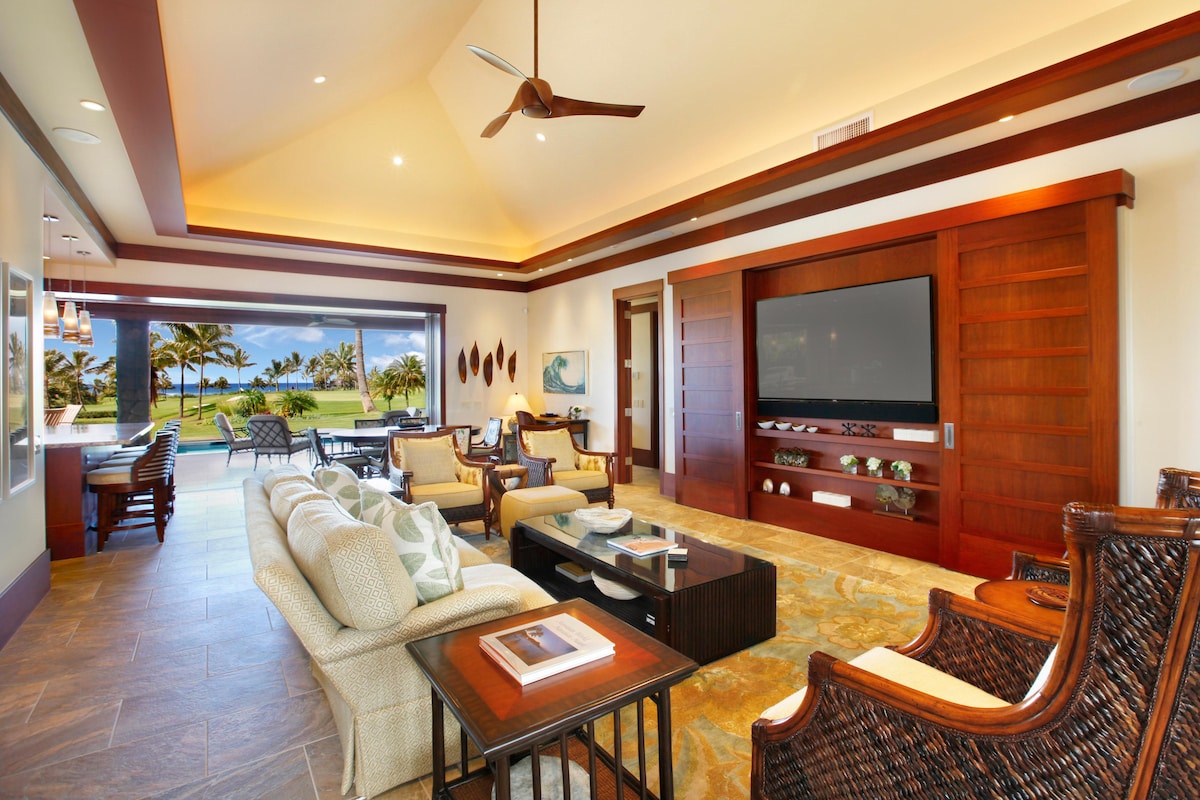 The living area displays an open floor plan with high ceilings and large glass doors leading to outdoor views. Comfortable seating is arranged around a central coffee table, and decorative elements include a large flat-screen TV mounted on a wooden wall feature.