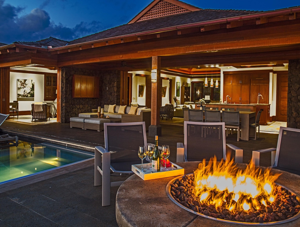 An outdoor seating area is depicted, featuring several lounge chairs arranged around a fire pit with vibrant flames. In the background, an open-concept interior reveals a modern kitchen and living space, illuminated by warm lighting, while the private pool and patio create a relaxing atmosphere.