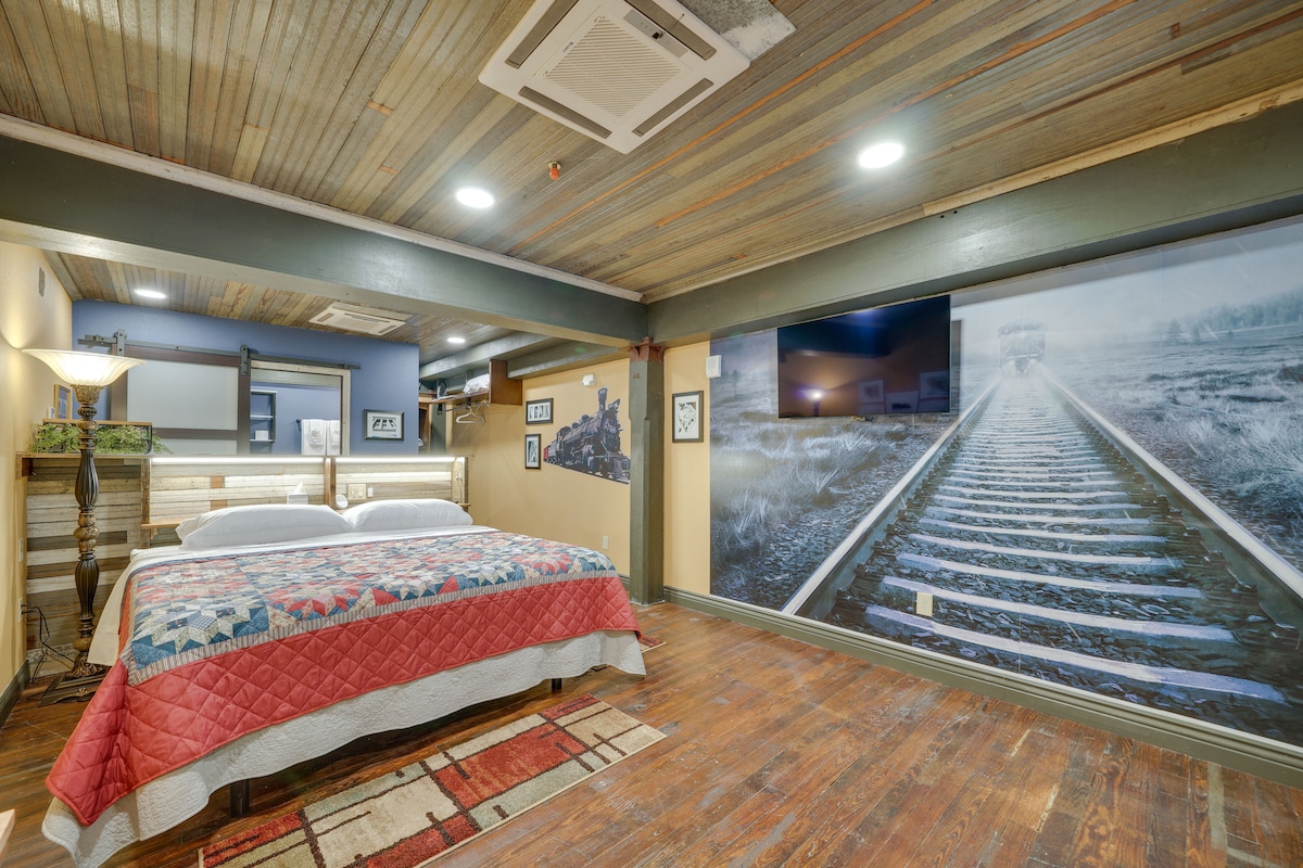 An open-concept studio features a spacious bed adorned with a patterned quilt. A large flat-screen TV is mounted on the wall opposite a mural depicting railroad tracks. Warm wooden flooring and soft lighting create a relaxed atmosphere.