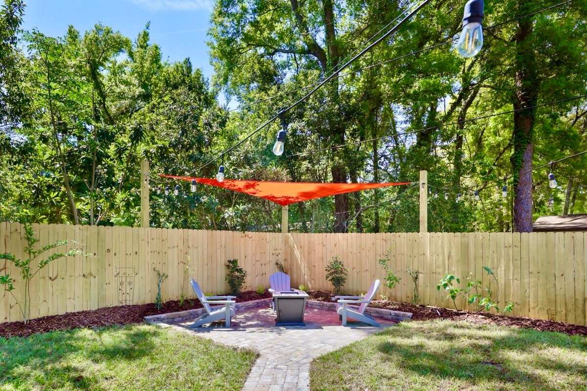 An outdoor recreation area features a fire pit surrounded by four adirondack chairs. Red shade sails provide coverage while string lights hang overhead. A wooden fence offers privacy, and greenery adds a natural touch to the serene setting.