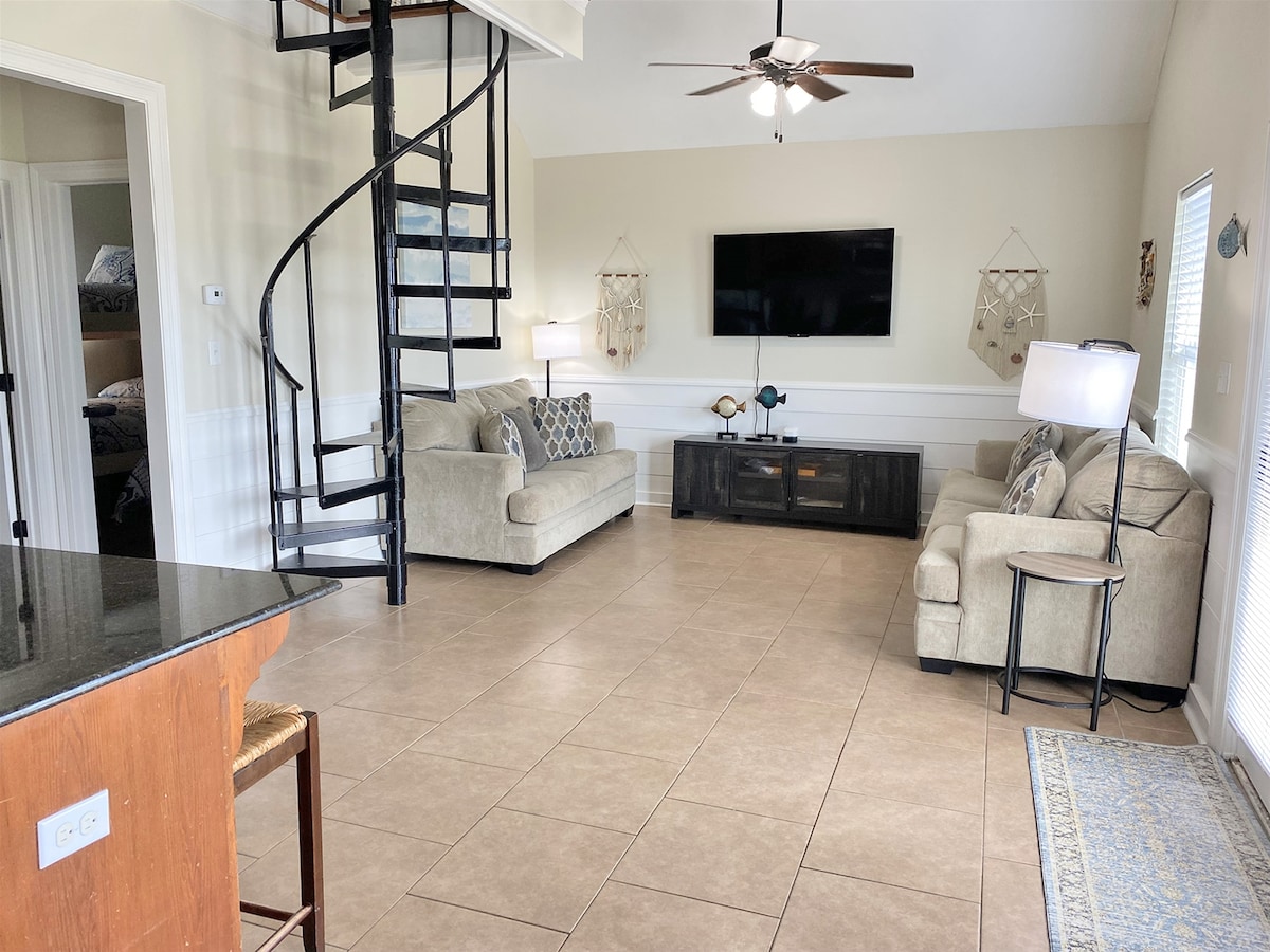 A light-filled living area is presented with a spiral staircase leading to the loft. Two neutral-toned sofas are arranged around a dark wood entertainment unit, which holds a flat-screen TV. Natural light streams through the nearby windows, illuminating the tiled floor.
