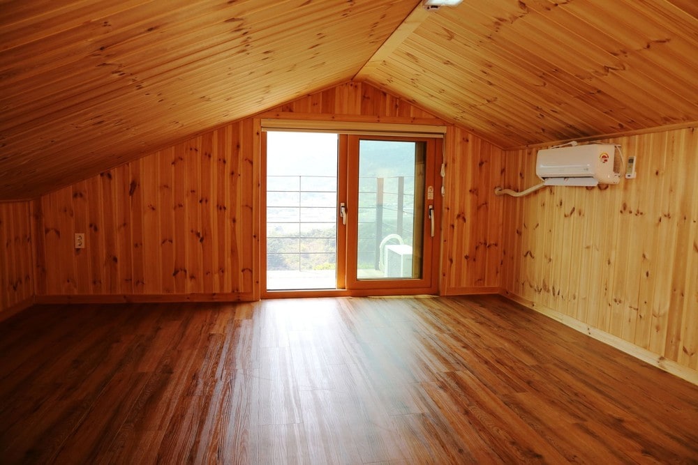 The attic space features warm wooden paneling and a sloped ceiling. Natural light floods in through a glass door, which opens to a balcony offering scenic views. Flooring is made of polished wood, contributing to the cozy atmosphere.