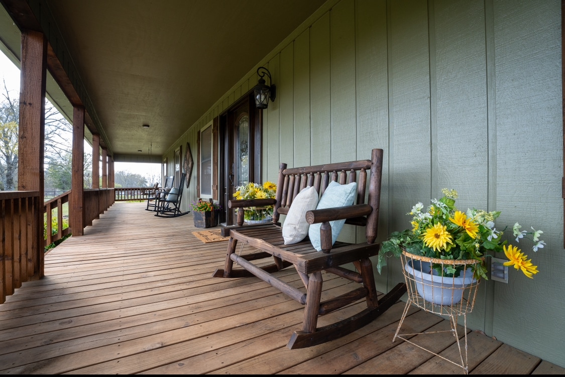 A spacious porch features a wooden rocking chair and a rustic bench adorned with soft pillows. Bright yellow flowers are arranged in a planter, adding a touch of color to the wooden flooring. The area is well-lit and invites relaxation.