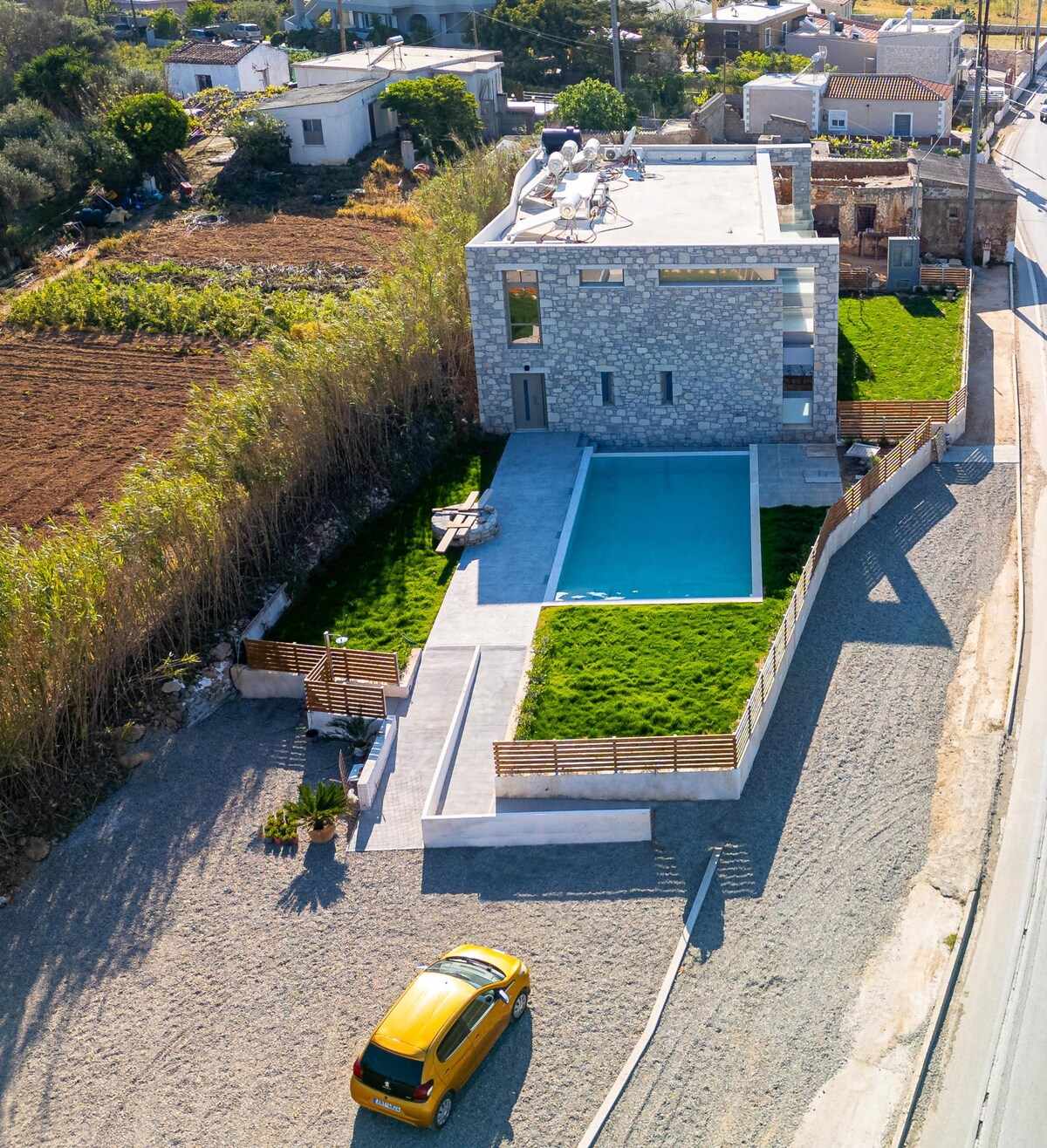 The aerial view captures a modern complex surrounded by greenery, featuring a shared pool and landscaped areas. A yellow car is parked nearby, while the building showcases a stone façade and clean lines. The surrounding land highlights a blend of cultivated fields and natural landscapes.