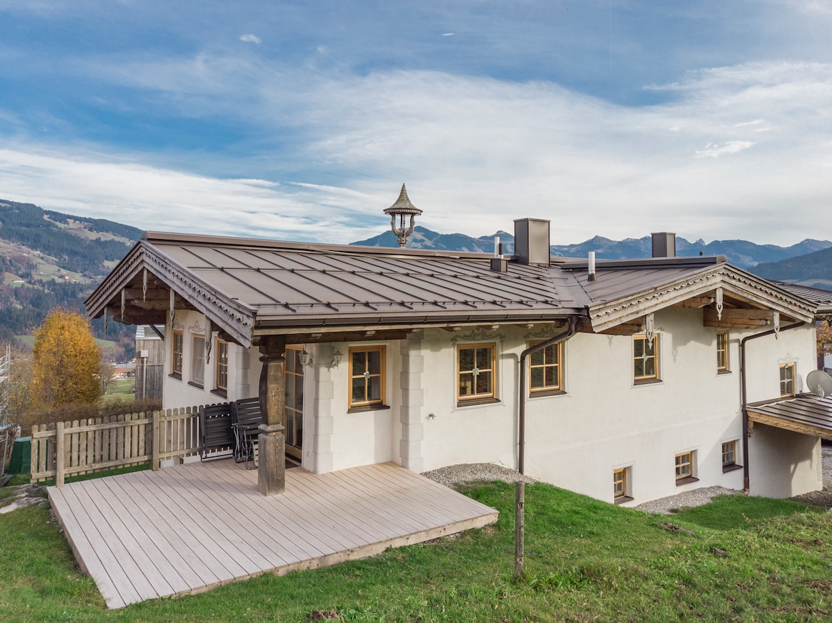 A spacious exterior view of the apartment is presented, showcasing a well-maintained wooden deck and a sloping green lawn. The charming structure features a brown roof and is surrounded by picturesque mountain scenery in the background, creating a serene atmosphere.