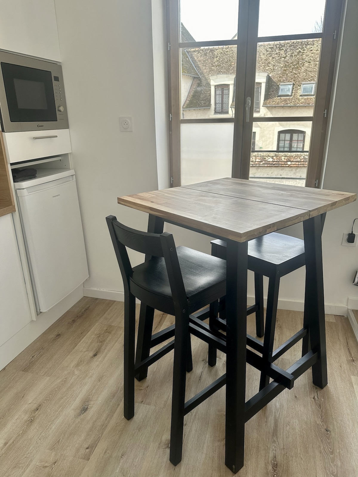 A compact kitchenette features a tall wooden table paired with two black stools. A microwave is integrated into the cabinetry, while a window allows natural light to enhance the space, offering a glimpse of the exterior views.