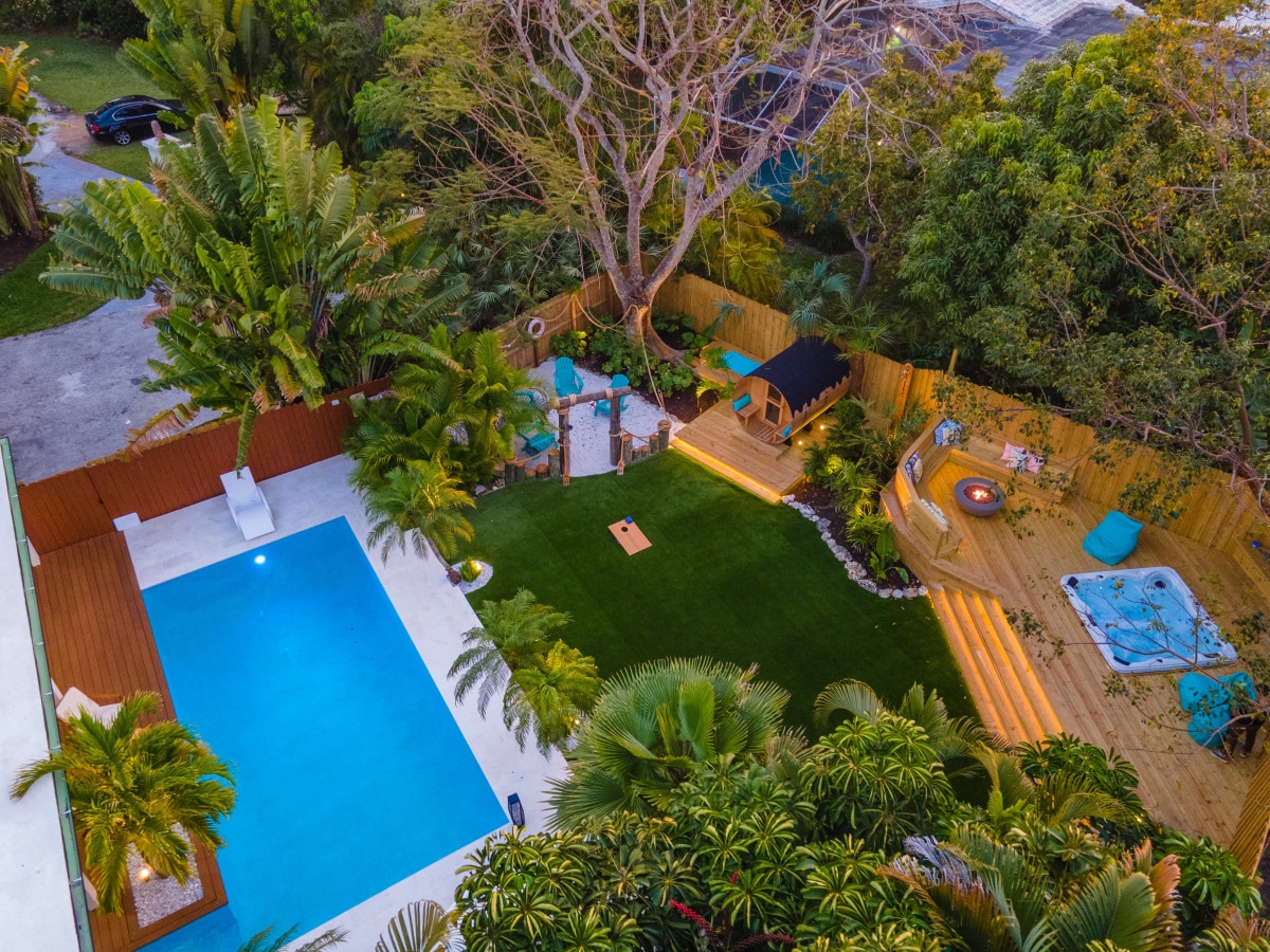 The outdoor area features a large blue swimming pool surrounded by tropical plants. A wooden deck offers a spa area with a jacuzzi, and a fire pit is located amidst a grassy area. Comfortable seating is arranged for relaxation and gathering.