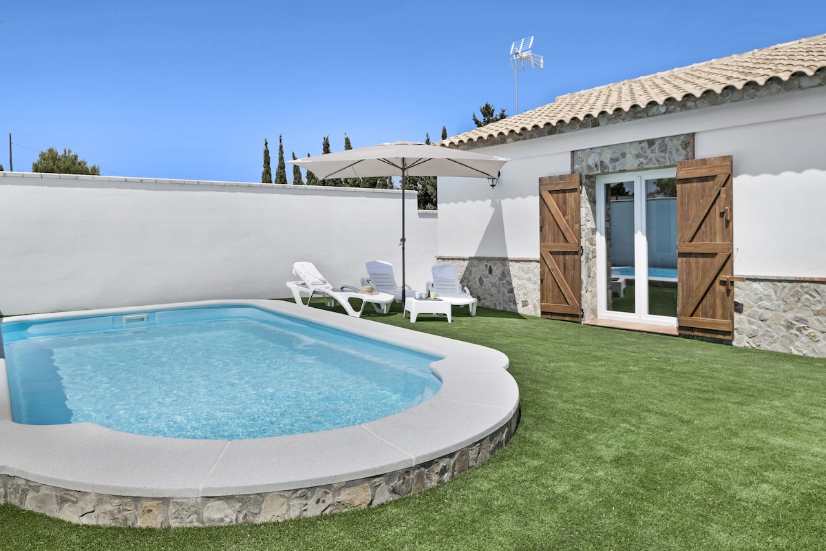 A private pool is framed by a well-maintained lawn, surrounded by white walls. Sun loungers are positioned nearby, and a large umbrella provides shade. Double doors lead from the pool area into the interior, enhancing the flow between indoor and outdoor spaces.