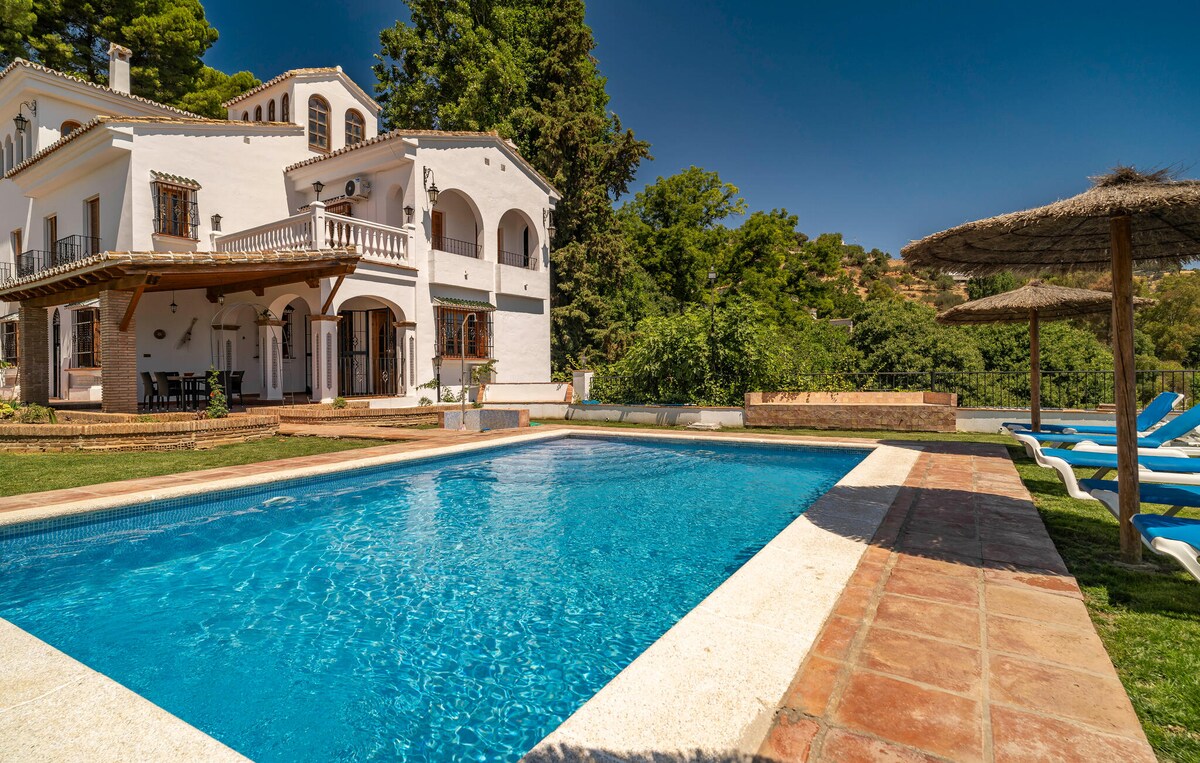 A spacious outdoor area features a refreshing swimming pool surrounded by green grass. Sun loungers are positioned along the pool's edge, with a thatched umbrella providing shade. The villa, with its white façade and terrace, is showcased in the background, framed by lush trees.