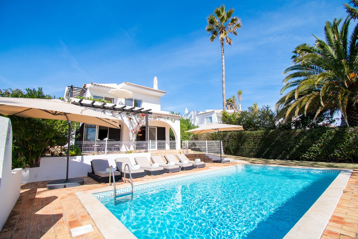 A private outdoor area features a clear swimming pool surrounded by sun loungers and shaded by large umbrellas. Lush palm trees provide a tropical backdrop. The villa's white exterior is visible in the distance under a bright blue sky.