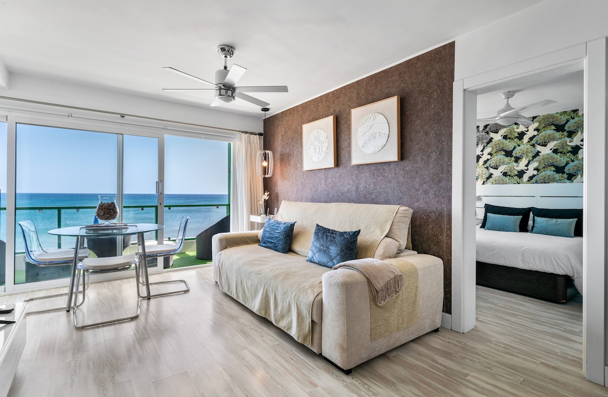 A living-dining area features a spacious sofa and a glass dining table with chairs. Large sliding doors provide a view of the Atlantic Ocean. The adjoining space includes a bedroom with patterned wallpaper and a large bed visible through an open doorway.