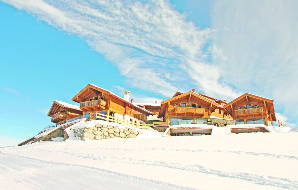 A group of luxurious chalets is set against a clear blue sky, surrounded by snow-covered ground. Each structure features wooden elements and large windows, while the sloped rooftops add to the chalet aesthetic. The landscape reflects tranquility and a connection to nature.