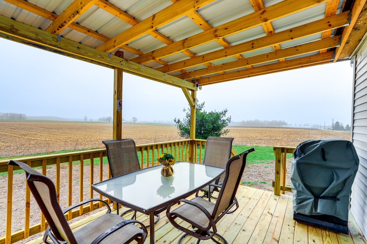 An outdoor deck area features a table with four chairs, offering space for dining. A gas grill is positioned nearby. The expansive field beyond the deck creates a serene rural backdrop, complemented by a low-hanging cloud cover.
