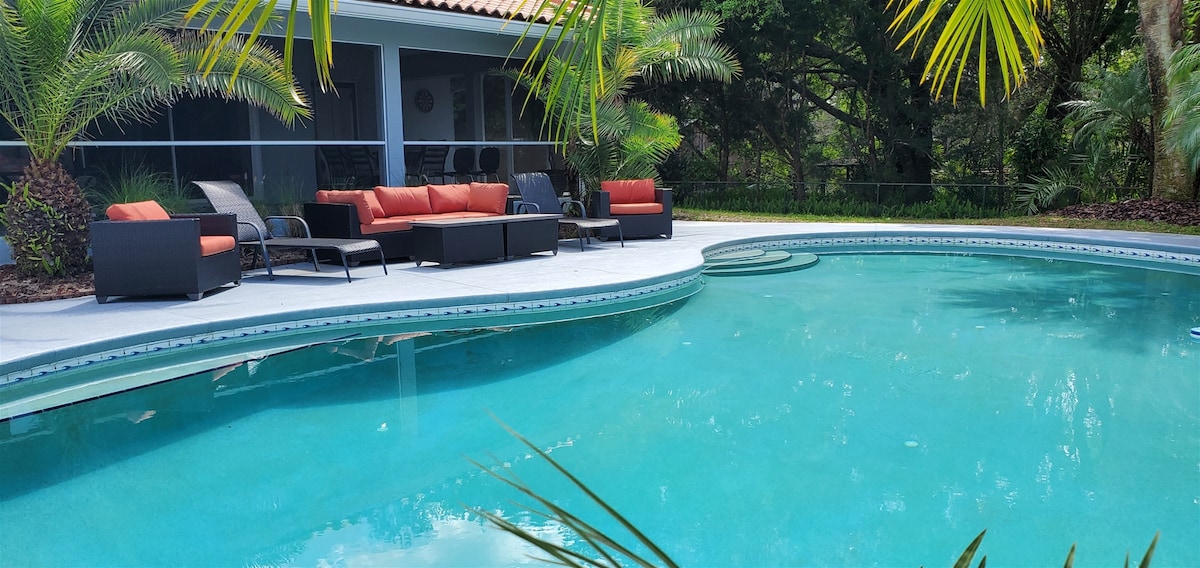 A serene infinity pool is surrounded by tropical plants, with comfortable outdoor seating and a coffee table positioned nearby. The pool's edge features a gentle curve, and the patio allows for easy access to the shaded lanai behind it.