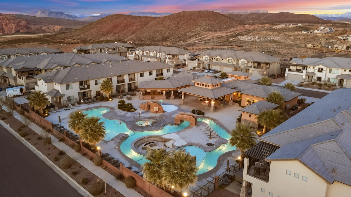 An aerial view of Zion Village Resort showcases the expansive pool area, featuring a lazy river and hot tub. Surrounding palm trees enhance the scenery, while the majestic red mountains stand in the background under a colorful sunset sky.
