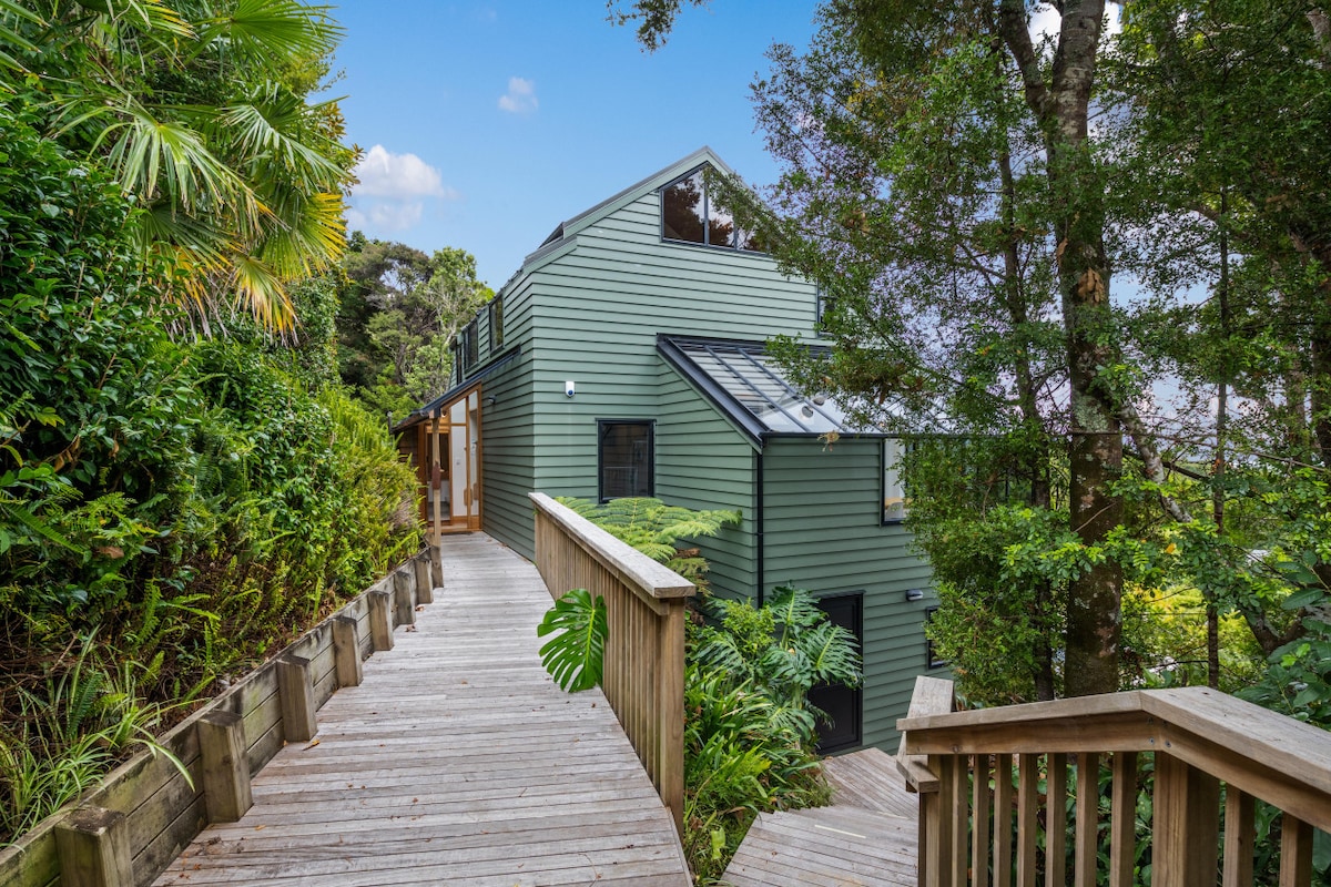 A pathway constructed of wooden boards leads up to a modern green house nestled among lush greenery. The exterior features large windows and a sloped roof, blending harmoniously with the surrounding foliage. Soft natural light enhances the tranquil atmosphere of the area.