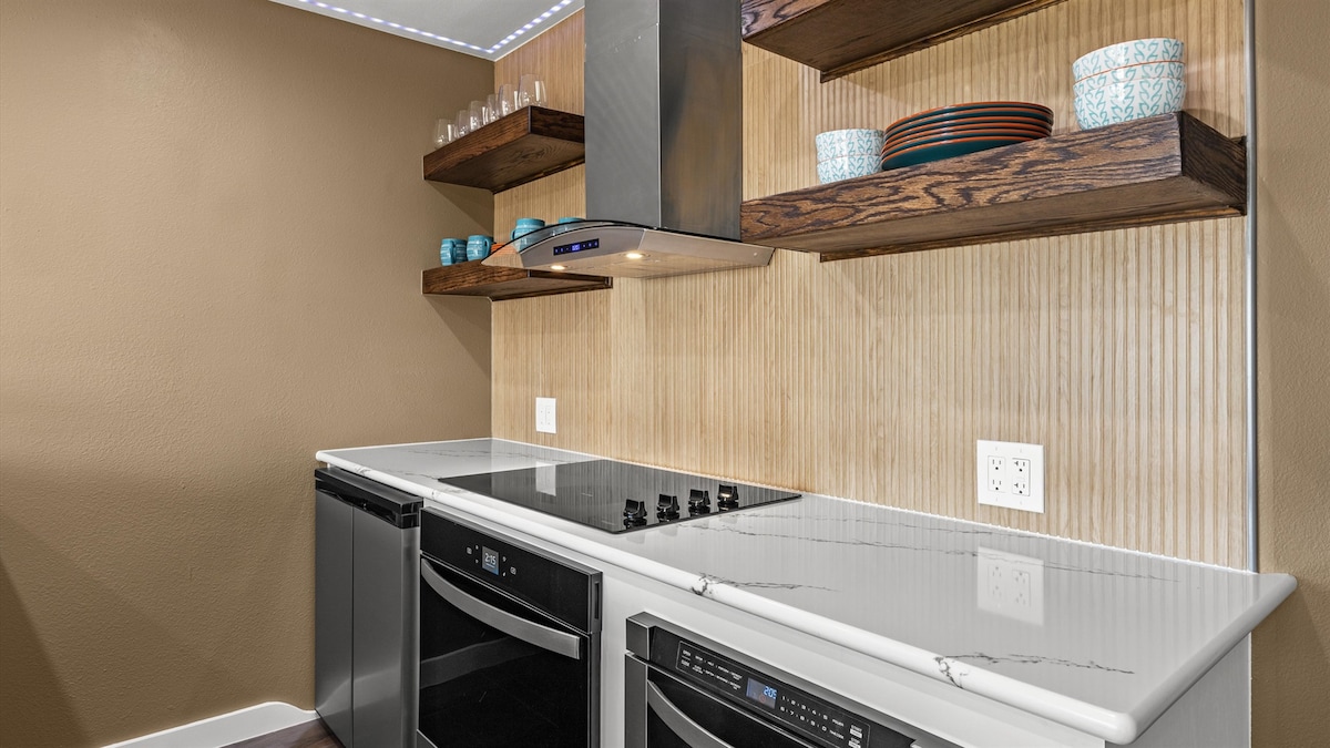 A modern kitchen features sleek countertops and contemporary appliances. Open shelves display colorful dinnerware, providing an organized and inviting look. The minimalist design is enhanced by under-cabinet lighting, illuminating the cooking area.