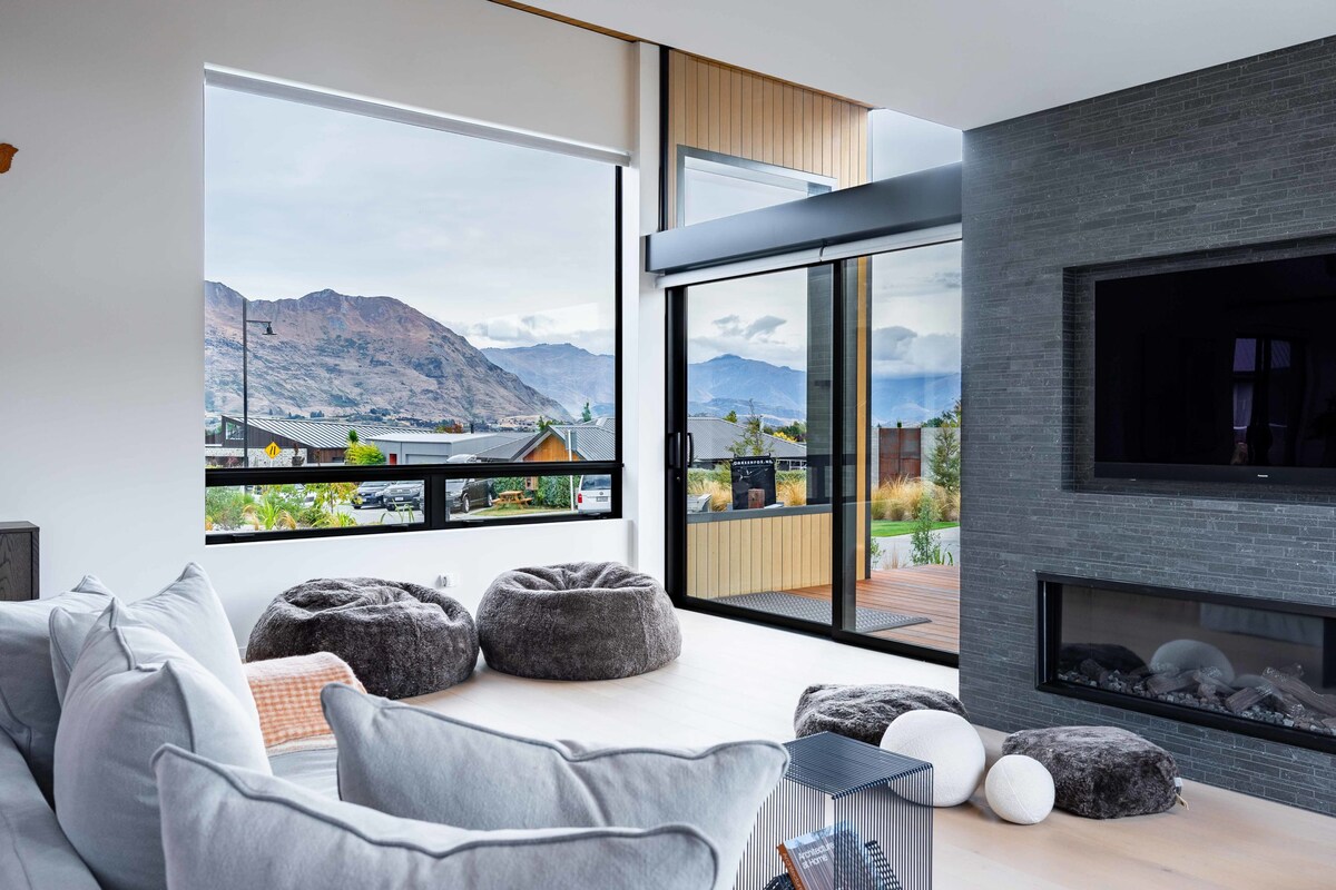 A modern living area features a comfortable sectional sofa and several bean bags arranged on light wood flooring. Large windows provide expansive views of the mountains, while a sleek wall-mounted TV and a contemporary gas fireplace contribute to the contemporary design.