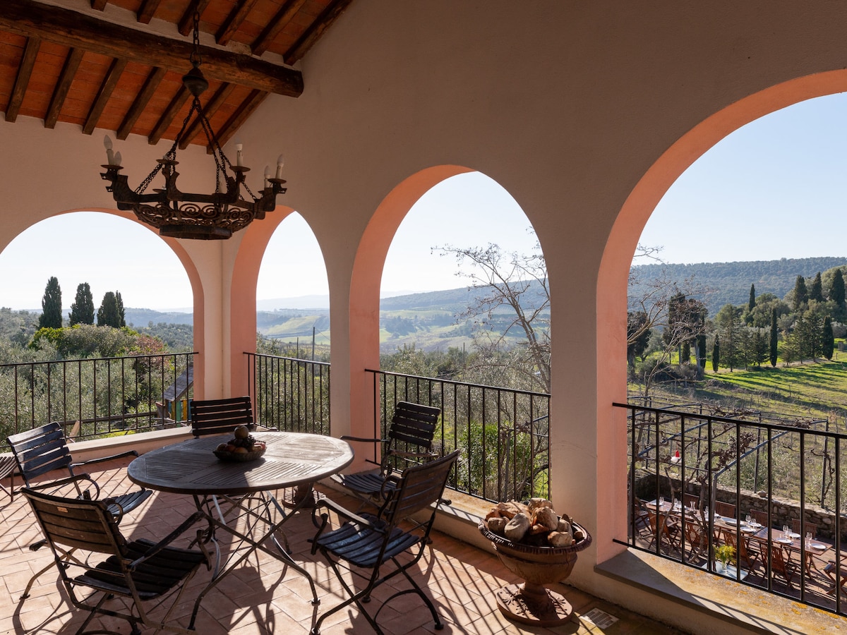 A terrace is framed by three arched openings, providing an expansive view of the rolling countryside. A round table with chairs is positioned in the center, enhancing the area for outdoor dining. The rustic charm is complemented by a combination of natural light and the surrounding greenery.