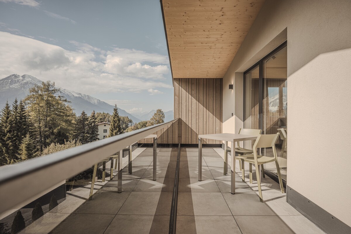 A spacious balcony features a sleek railing and a wooden ceiling, offering views of surrounding mountains and trees. Two light-colored chairs and a table are positioned to create an inviting outdoor space for relaxation and enjoyment of the natural scenery.