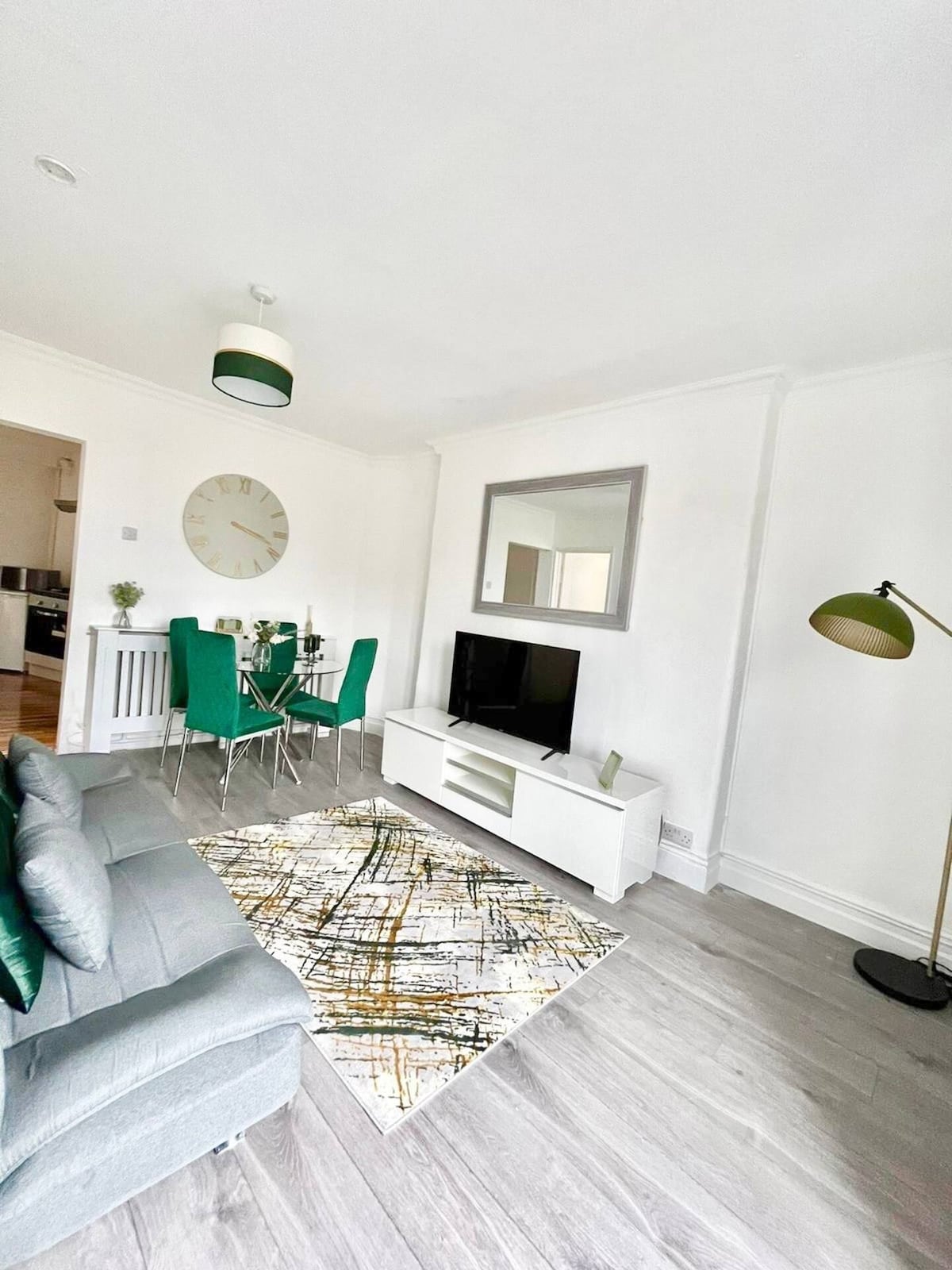 A modern living area features a spacious grey sofa and an elegant TV unit with a 43-inch flat-screen TV. A circular wall clock and a large mirror add depth to the bright space. A dining area is visible, with a table and green-accented chairs beside a window.