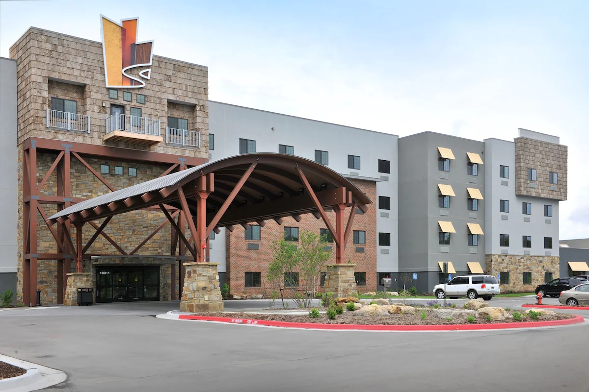 The hotel exterior features a modern rustic design, with a prominent stone façade and wooden beams. The entrance is marked by a spacious overhang, leading to a glass double door entrance. Surrounding landscaping includes rocks and low shrubs, creating an inviting atmosphere.