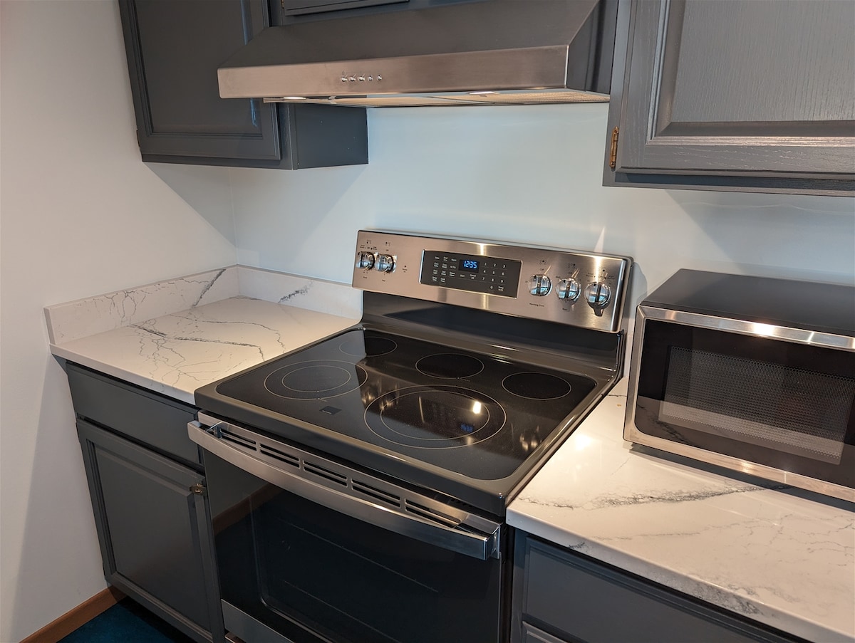 A modern kitchen area features a sleek stainless steel stove with an oven beneath and a glass induction cooktop. Gray cabinets complement the white marble countertops, alongside a compact microwave positioned nearby. A well-organized layout invites ease of use for cooking and meal preparation.