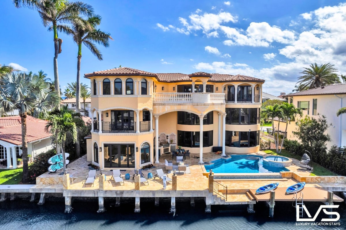 The exterior of a waterfront estate is showcased, featuring a generous pool area with multiple lounge chairs. A large terrace extends from the main structure, which features several rounded glass walls. The property is surrounded by palm trees and kayaks are visible along the dock.