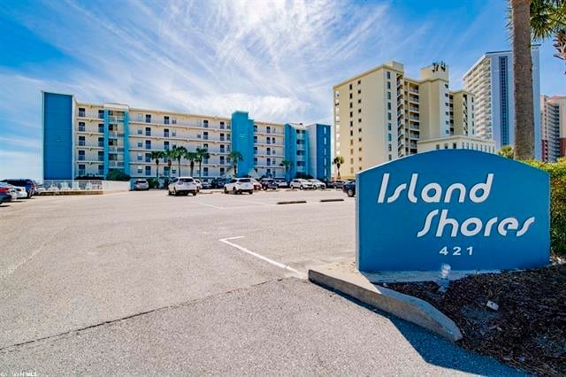 The exterior of Island Shores is depicted, featuring a modern six-story building in blue and white. The entrance sign displays the property name and number, surrounded by palm trees. The parking area accommodates several vehicles, emphasizing the spaciousness of the complex.