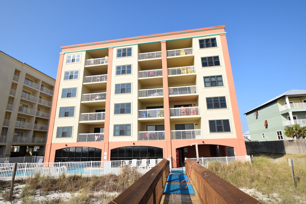A four-story beachfront condominium is depicted, featuring a bright coral and cream exterior. Balconies with glass railings are visible on each floor, providing access to outdoor spaces. A wooden walkway leads toward the pool area nestled among sandy surroundings, emphasizing the proximity to the beach.