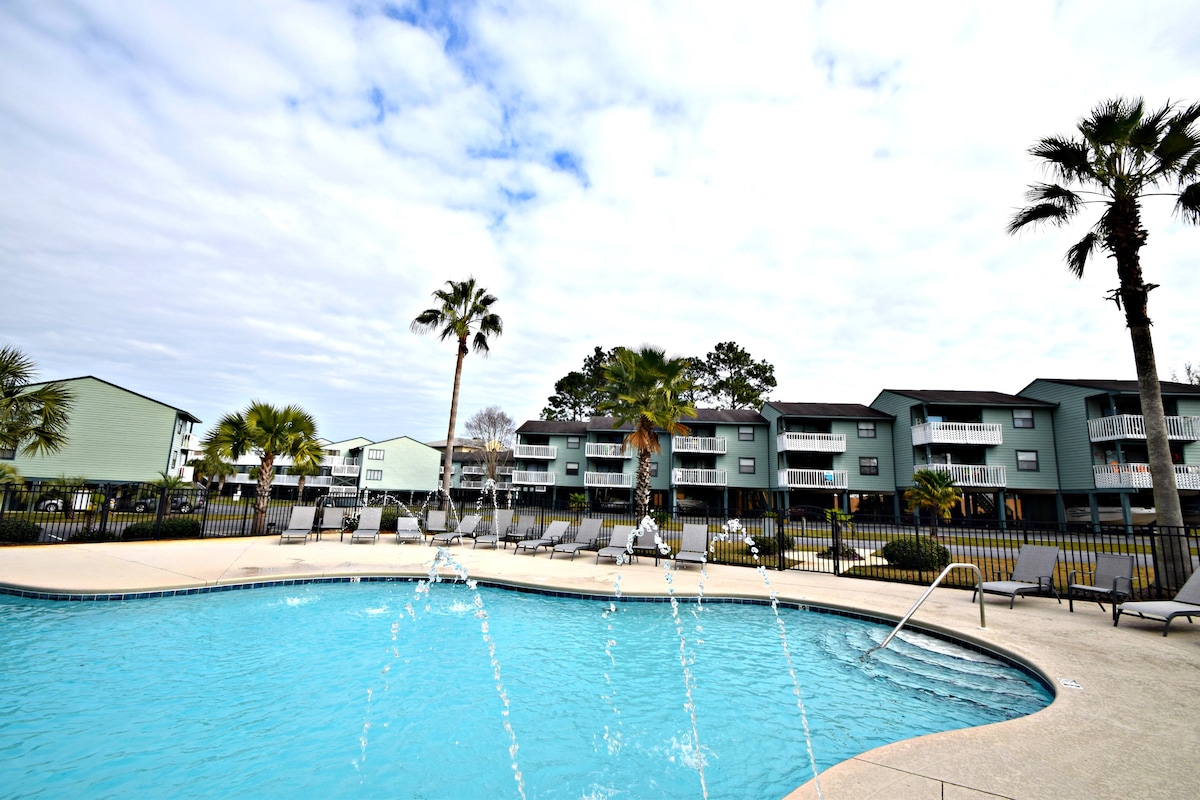 A sparkling pool is surrounded by lounge chairs, palm trees, and well-maintained landscaping. The tranquil water features are visible, providing a refreshing atmosphere. In the background, multiple two-story buildings showcase balconies, creating a welcoming environment for guests.