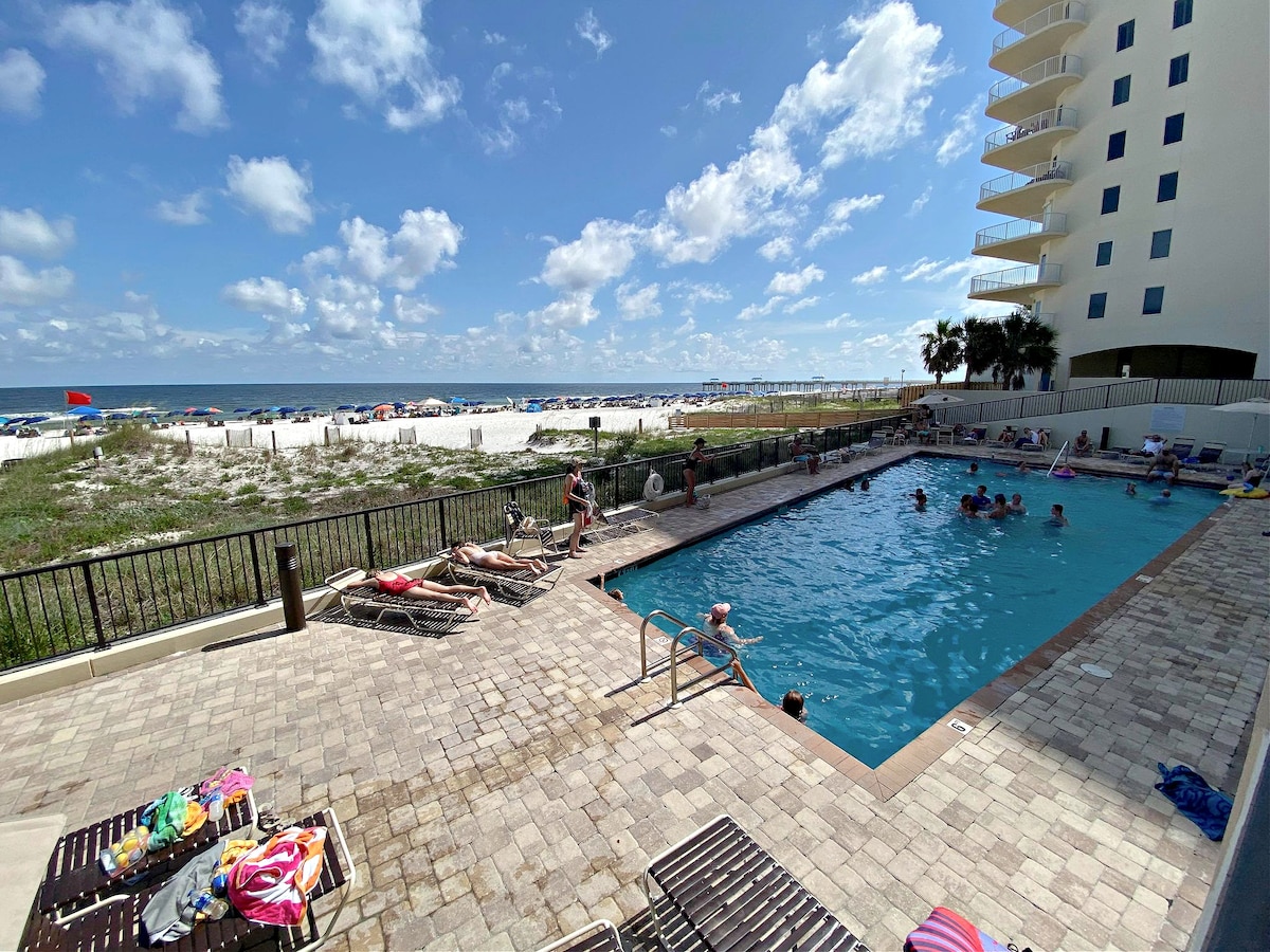 An outdoor pool is framed by lounging chairs situated on a paved deck, overlooking the beach. The clear blue water reflects the sky, and beach-goers can be seen relaxing in both the pool and on the sandy shore in the background.