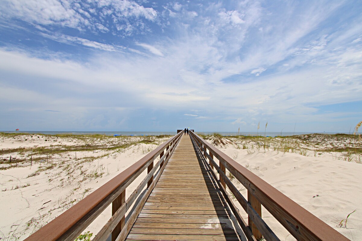 A wooden boardwalk stretches toward the beach, flanked by soft white sand and green grass. The expansive sky overhead displays a mix of clouds, enhancing a serene atmosphere. The distant view reveals the ocean meeting the horizon.