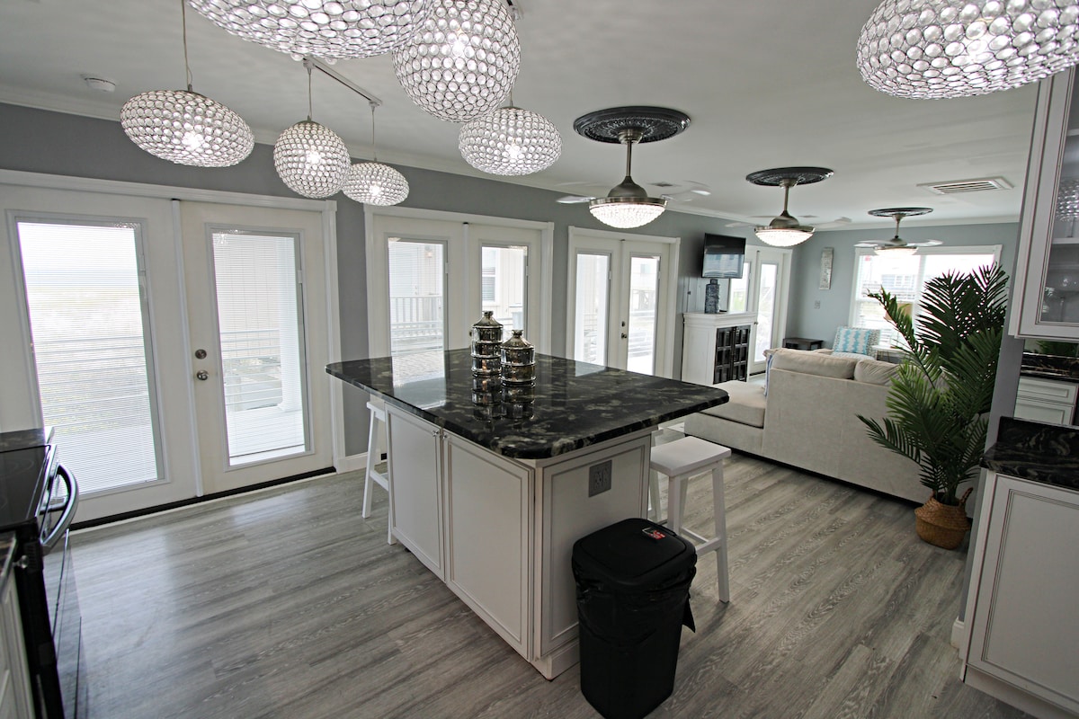 A modern kitchen is featured with a large dark countertop island at its center, surrounded by stylish bar stools. Elegant light fixtures hang from the ceiling, enhancing the bright space filled with soft colors. Large sliding glass doors offer views of the outdoor area, allowing ample natural light.