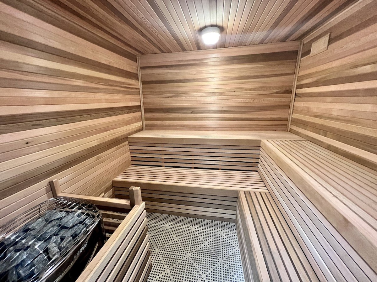 A wooden sauna interior is depicted, featuring light-toned wooden walls and bench seating arranged in tiers. The stone floor is gridded for drainage, and a metal container filled with sauna stones is visible against the wall. Soft lighting is provided by a ceiling fixture.