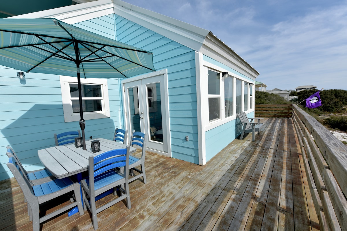 A sunlit outdoor deck features a dining table with blue and gray chairs, complemented by an umbrella for shade. Large windows allow natural light into the cottage. The deck overlooks lush greenery and the coastline, showcasing a beach atmosphere.