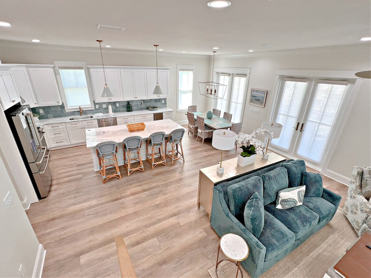 An open-concept kitchen and living area features light-colored cabinetry and a large island with bar seating. A cozy seating area with a blue sofa is visible, alongside a dining table and chairs. Light fills the space through multiple windows, creating an inviting environment.